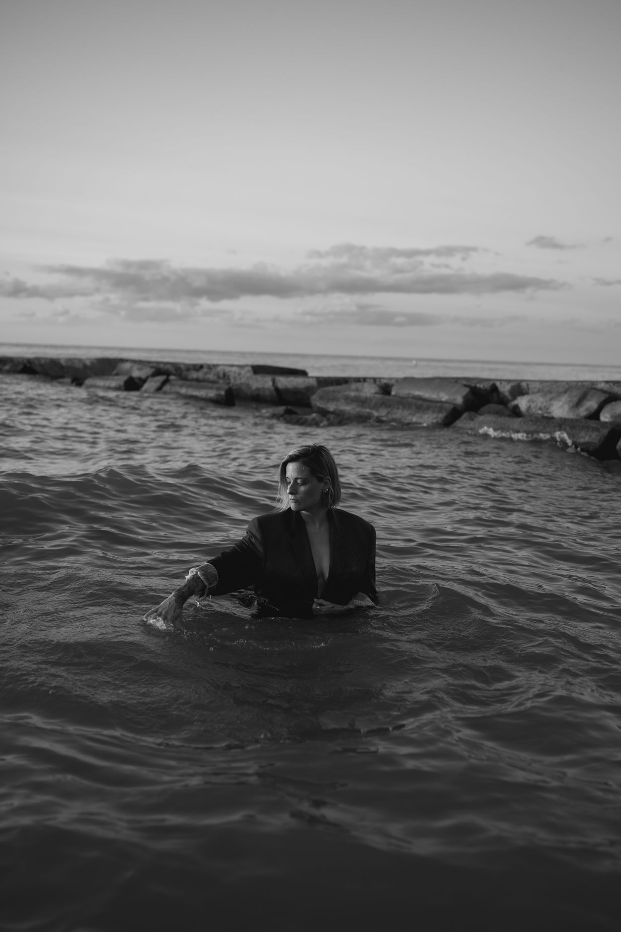 Meg in the water in a dark blazer, one arm lifted, breakwall on the horizon behind her.