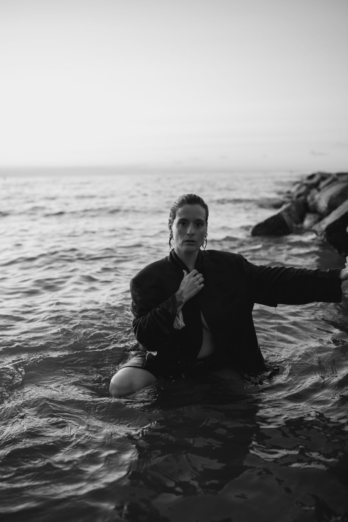 Meg kneeling in shallow water in a dark blazer, hand at her chest, looking directly at the camera.