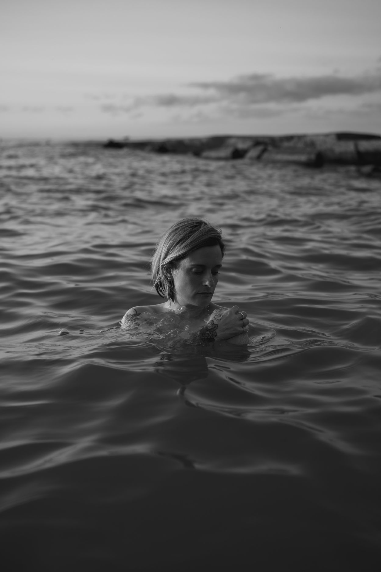 Meg submerged to the shoulders in still water, arms crossed over her chest, eyes lowered.